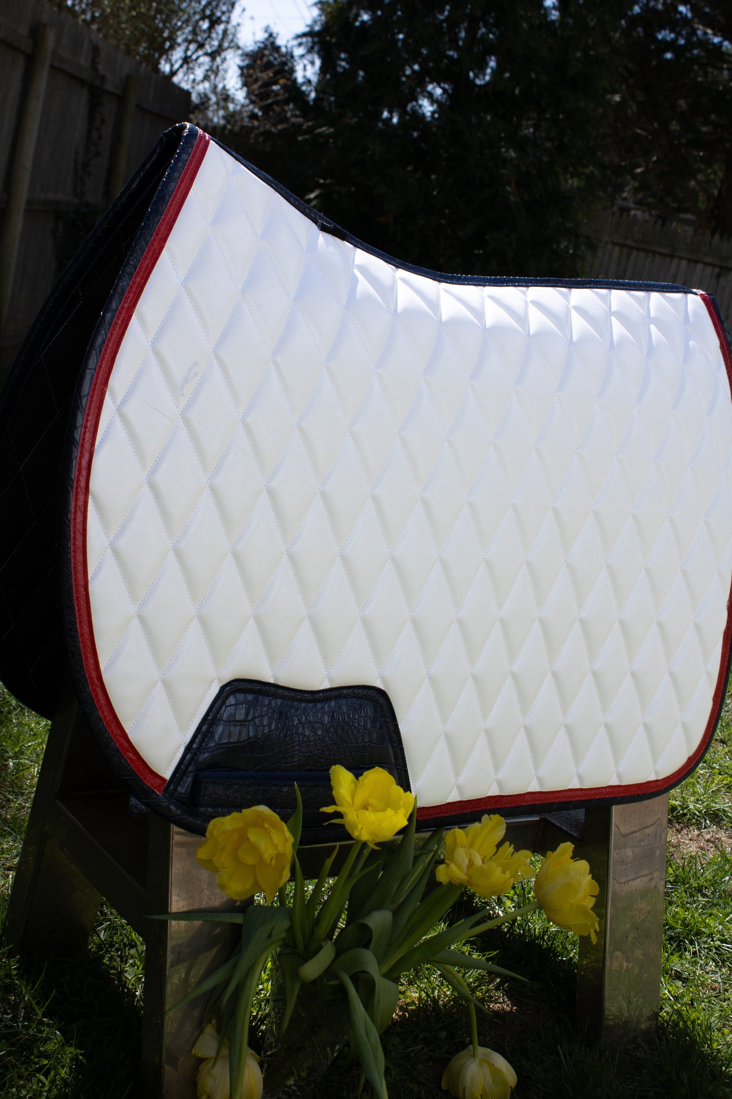White quilted saddle pad with red trim on a stand, surrounded by yellow flowers and greenery.
