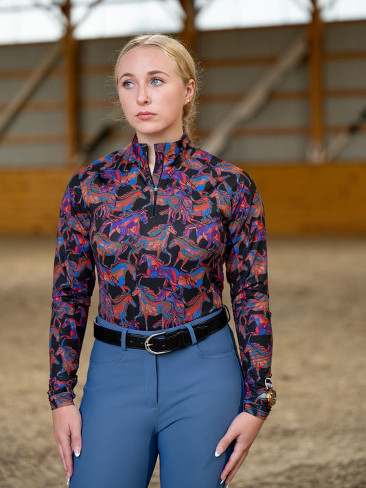 Woman wearing a patterned equestrian shirt and blue pants in an indoor arena.