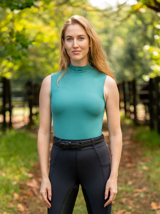 Woman wearing a teal sleeveless top and black pants standing outdoors with greenery in the background